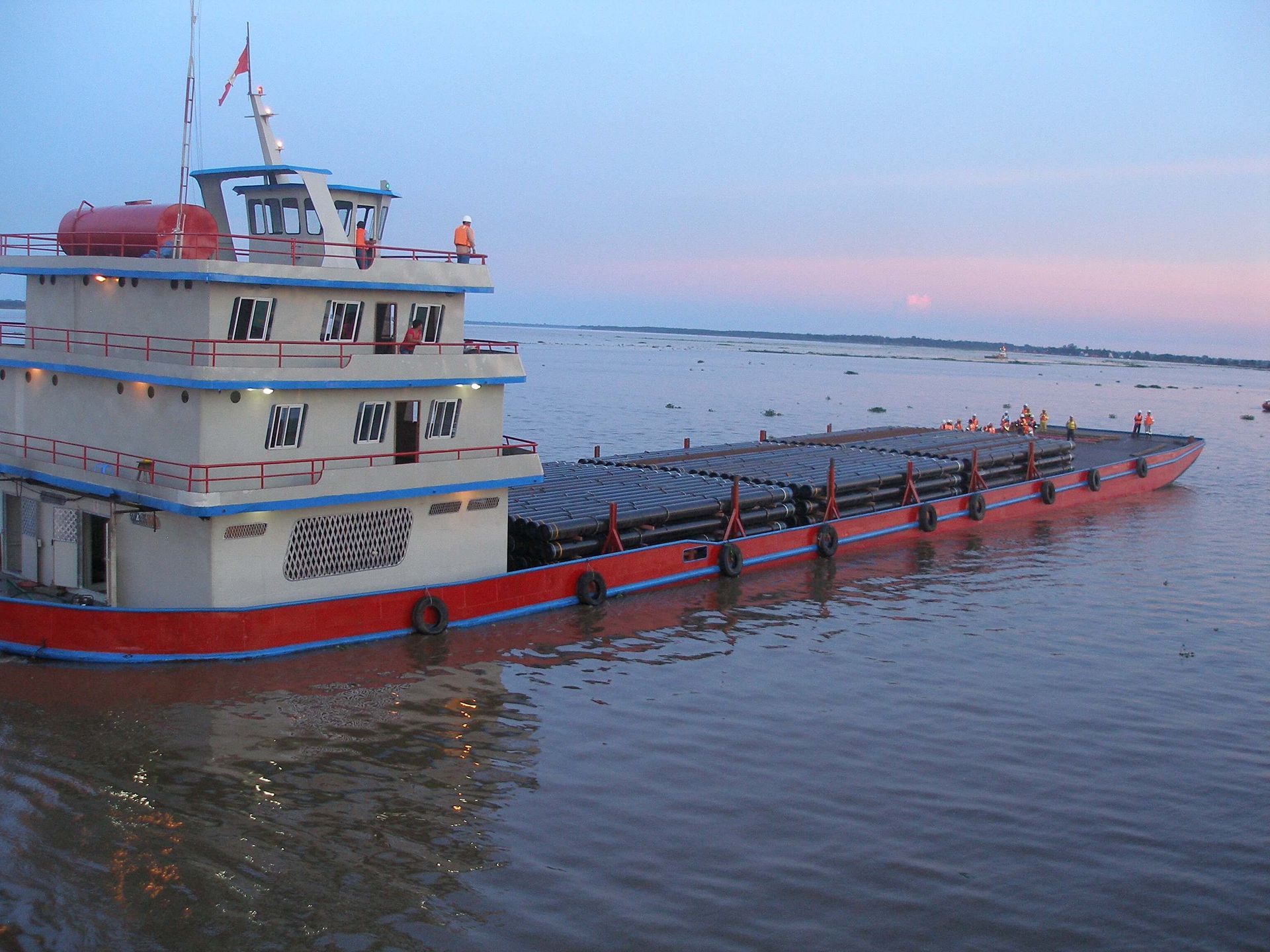Large red and white boat on water, carrying cargo, with a cloudy sky in the background.