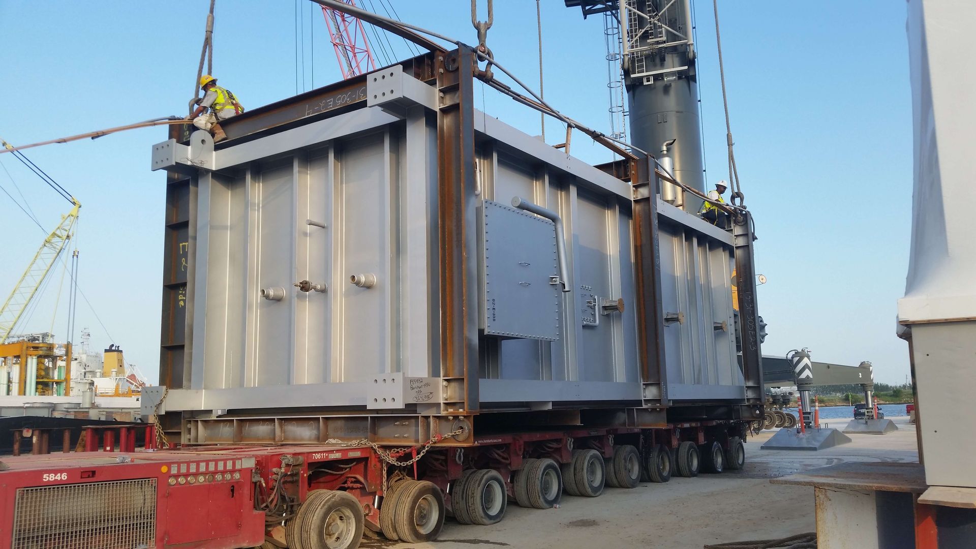 Large, gray metal container being lifted by a crane onto a red truck bed, outdoors.