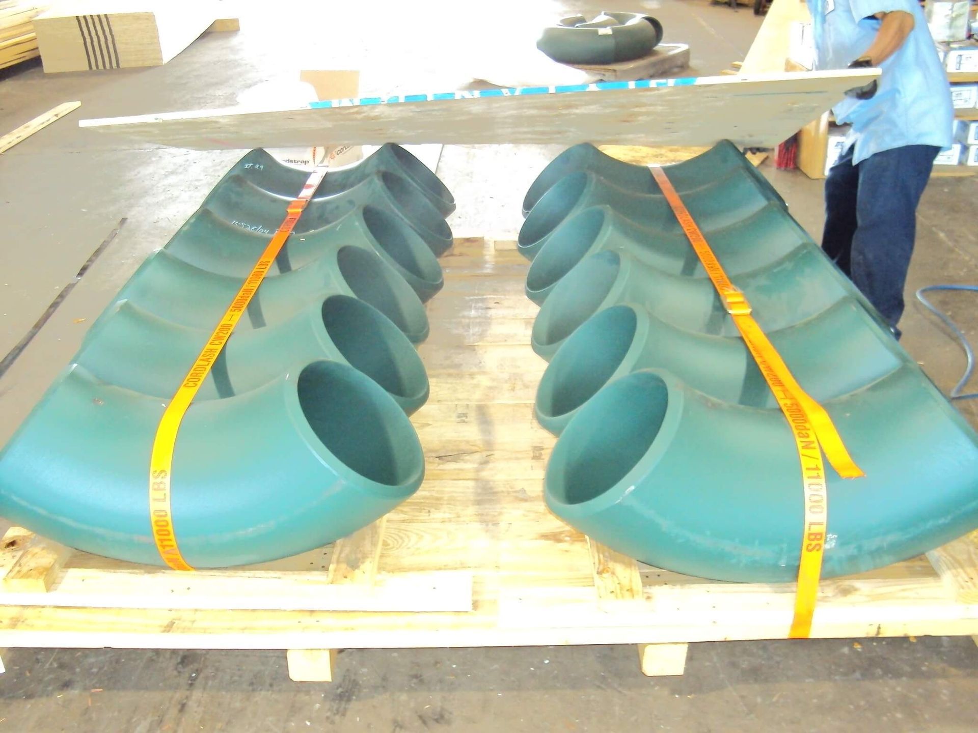 Two rows of large green pipe elbows secured on a pallet with yellow straps; a person loads boxes.