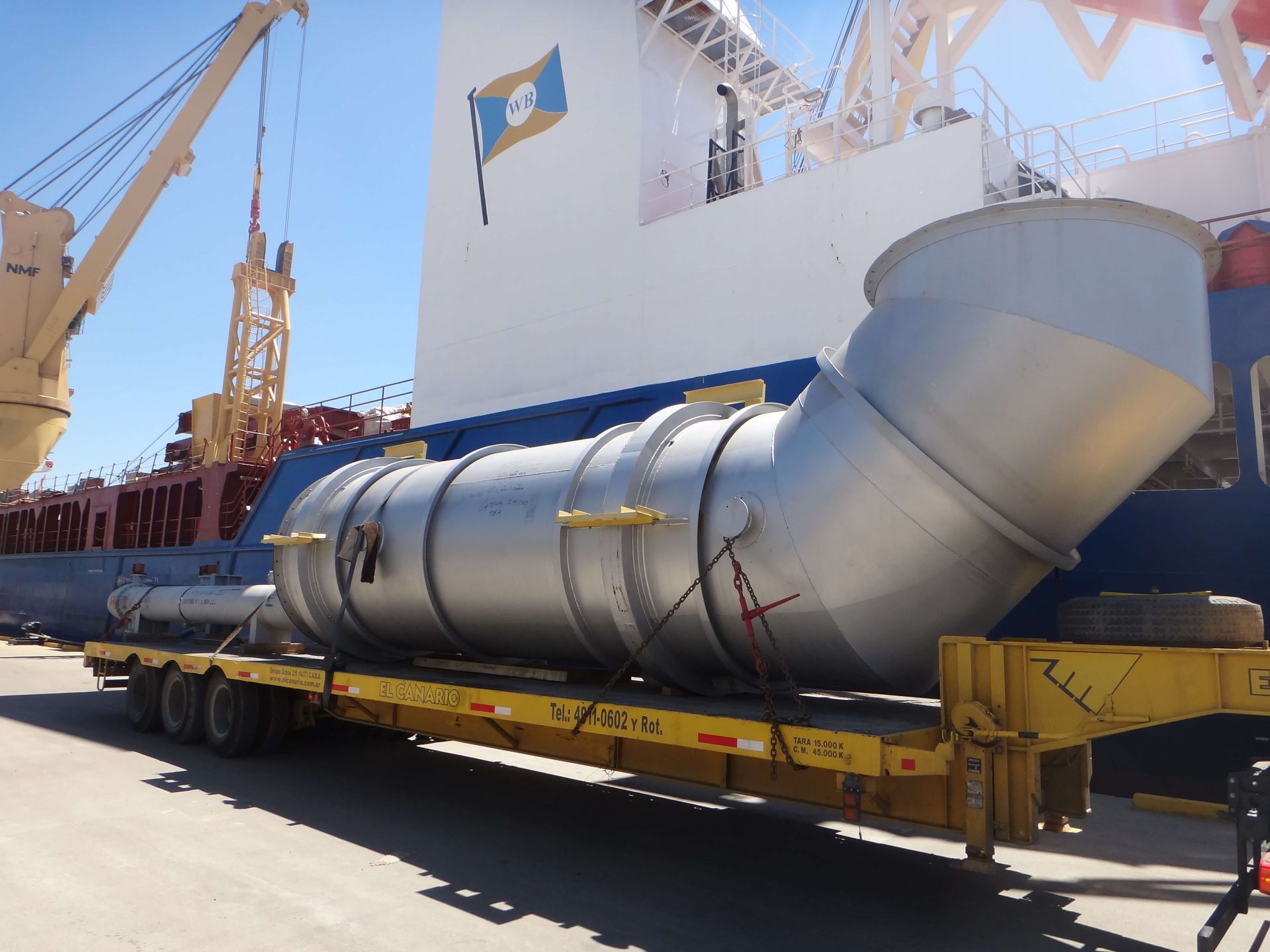 Large metal pipe section on a flatbed trailer next to a ship; blue sky.