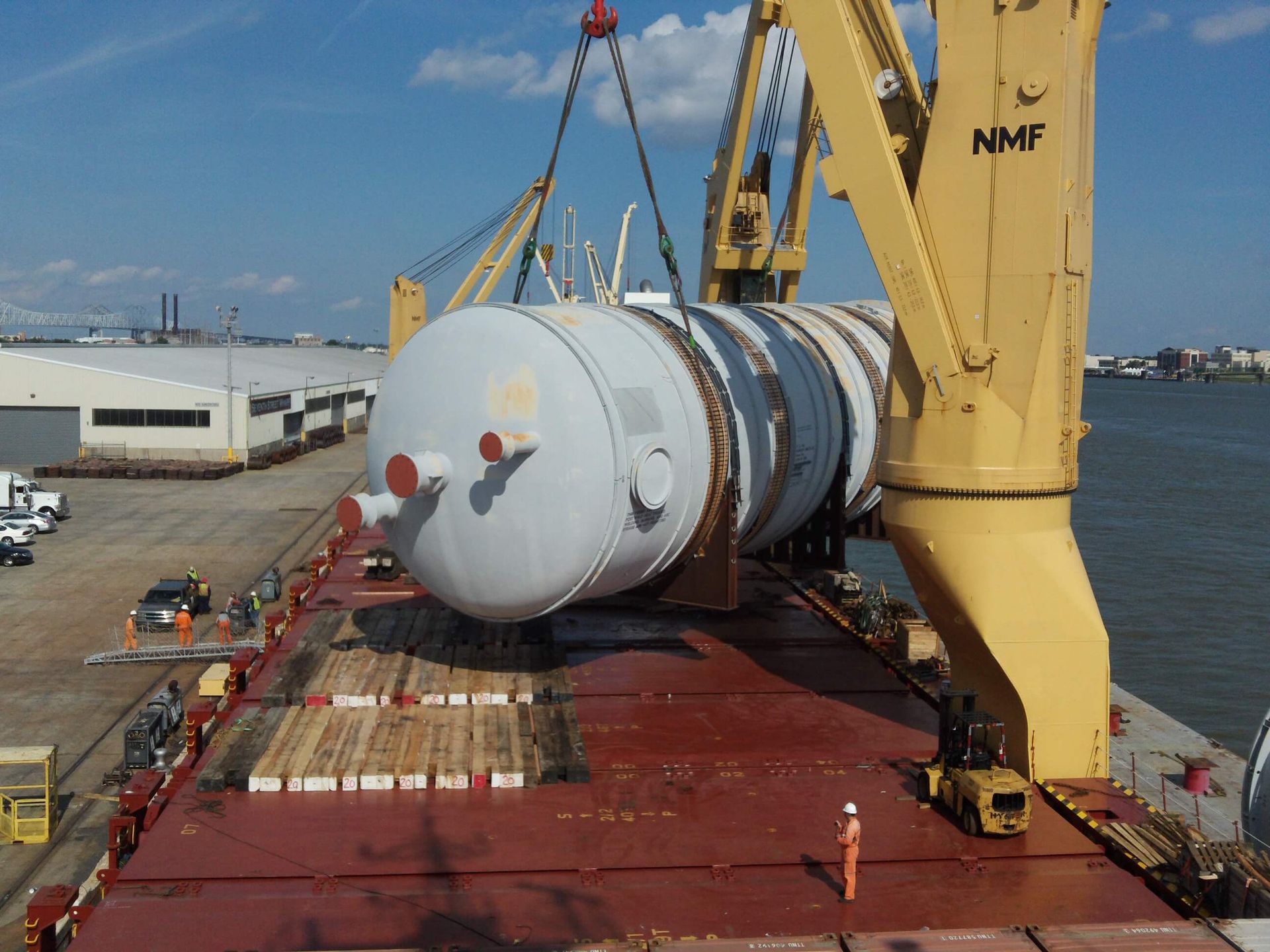 Large cylindrical tank being lifted by a yellow crane onto a cargo ship on a sunny day.