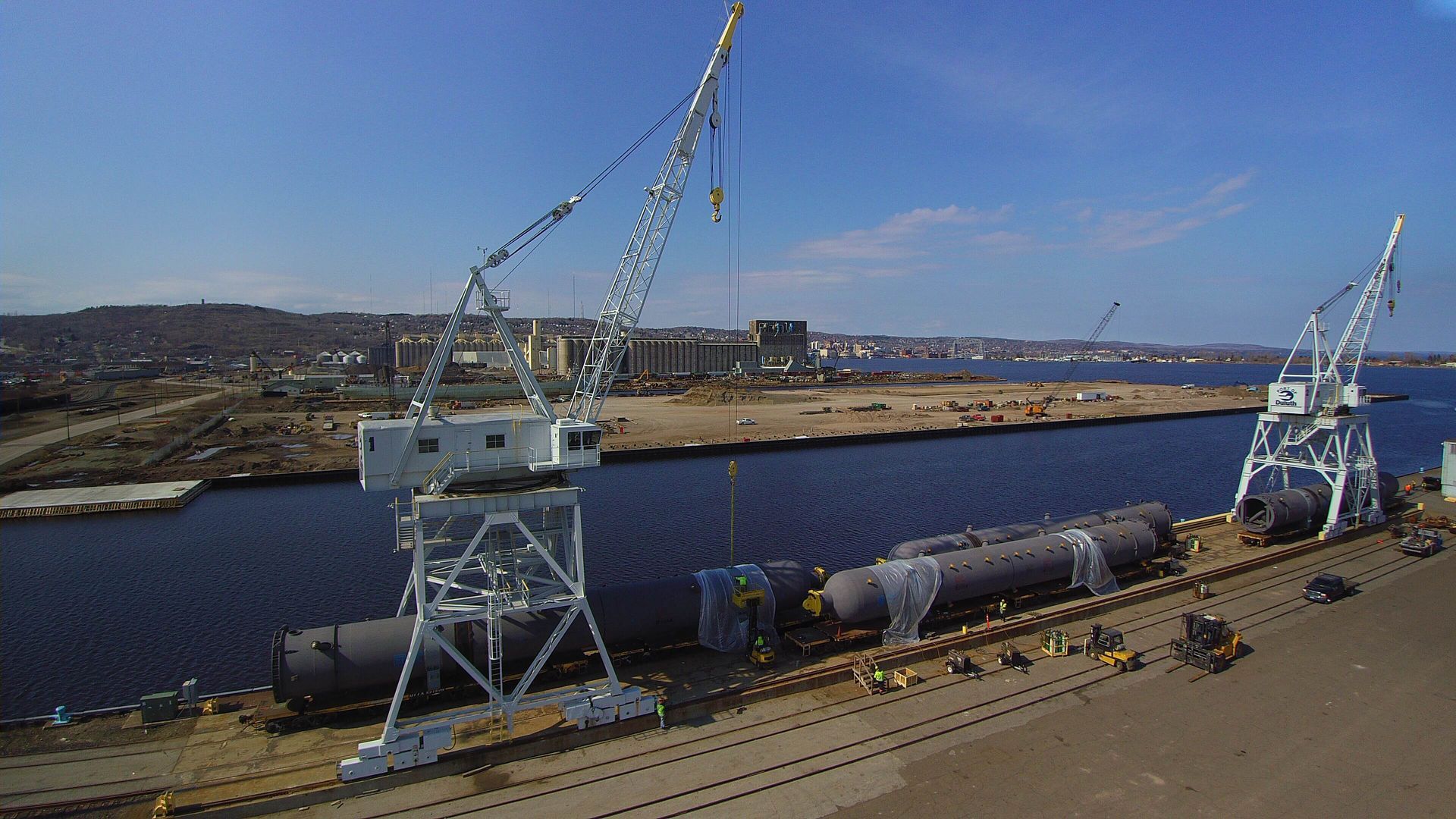 Two cranes on a dock loading large cylindrical objects. Industrial harbor with buildings and water in the background.