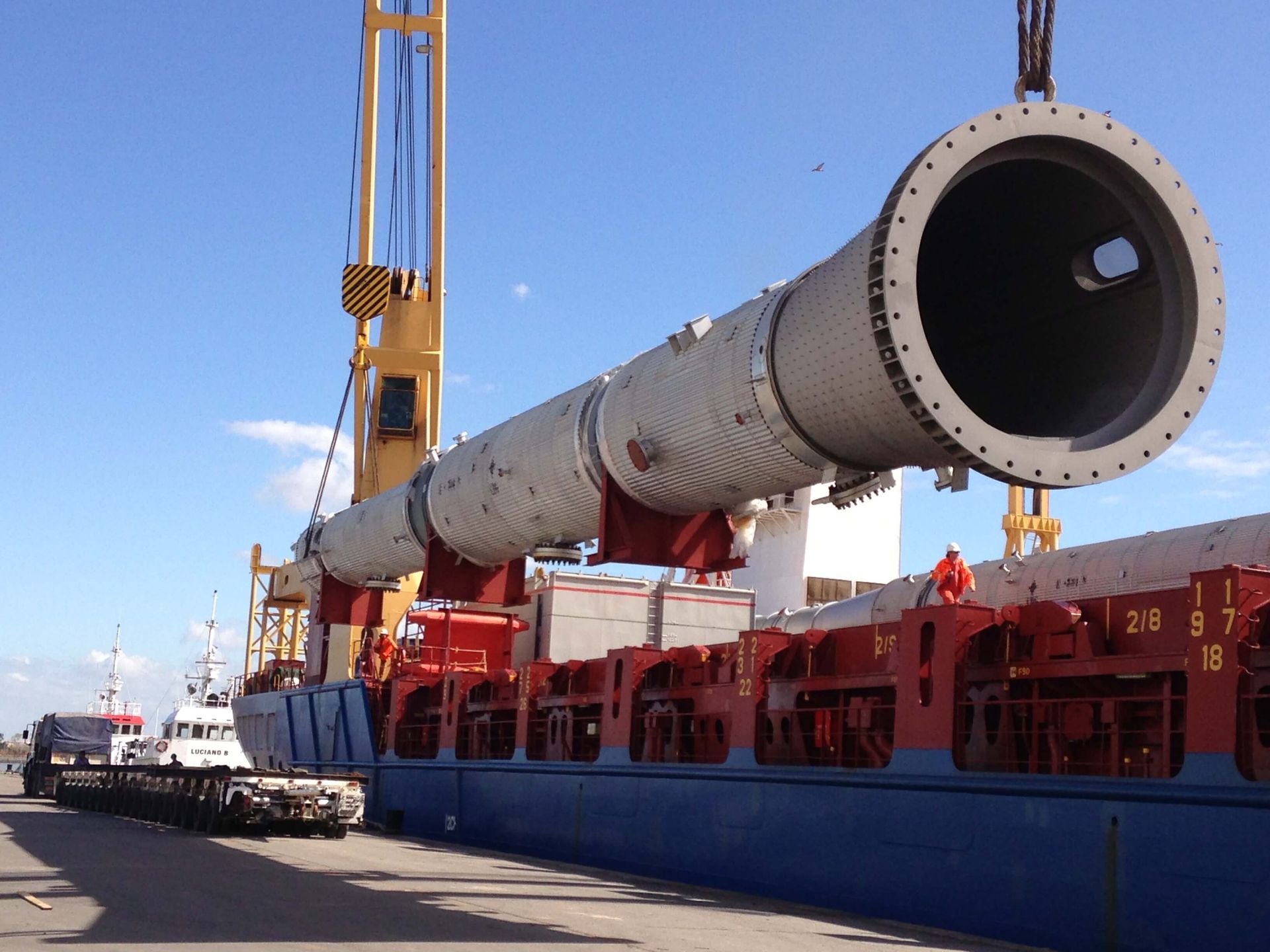 A large cylindrical industrial component being lifted by a crane onto a ship at a port.