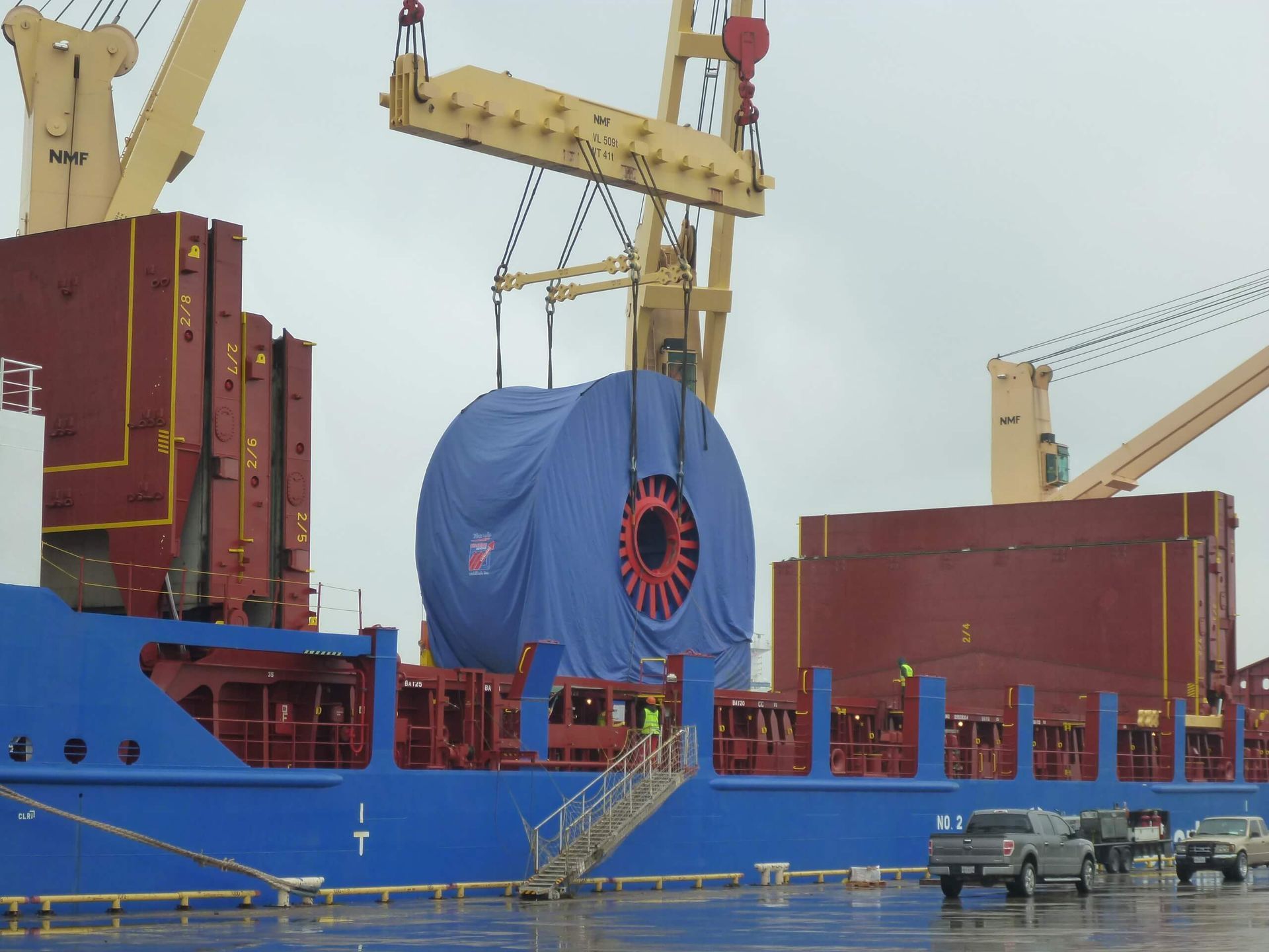 Large blue-wrapped spool being lifted by crane onto a blue ship; two trucks parked on a dock nearby.
