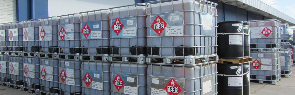 Tanks and barrels of chemicals stacked in a warehouse. Each is labeled with a hazard symbol.
