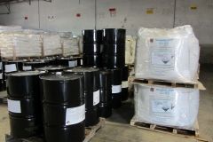 Black barrels and white bags of chemicals stacked on pallets inside a warehouse.