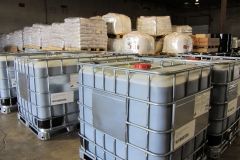 IBC totes filled with liquid, arranged in a warehouse. Pallets of white materials in the background.