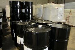 Black barrels stacked in a warehouse setting; some are sealed with white caps, while others are open.