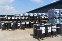 Rows of black barrels, possibly chemical storage, outside a building.  Blue sky and clouds in background.