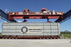 Large container being lifted by a red overhead crane; the container is on a multi-axle trailer.