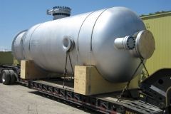 Large, cylindrical, silver tank secured on a flatbed trailer with wooden supports; outdoors.
