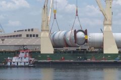 Large cylindrical industrial equipment being lifted off a ship by cranes. Tugboat nearby.