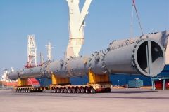 Large industrial pipes on a flatbed trailer at a port, being lifted by a crane.