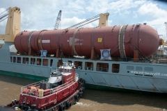 A large red industrial tank being transported on a ship, assisted by a red tugboat.