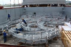 Workers assembling curved metal walkways inside a cargo ship, likely for a pier or terminal.
