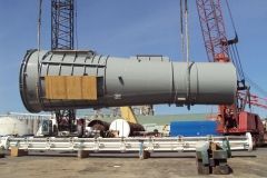 A large gray cylindrical structure being lifted by cranes at a construction site, blue sky in background.