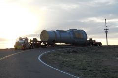 A large, cylindrical industrial component on a flatbed truck rounding a rural road at sunset.
