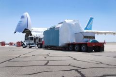 A large cargo plane unloading a large, wrapped container from a flatbed trailer at an airfield.