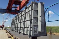 Large metal flood gate being lifted by a crane near water, with blue sky and bridge in background.