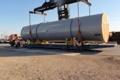 Large cylindrical tank suspended by a crane, being loaded onto a flatbed trailer outdoors. Workers in safety vests present.
