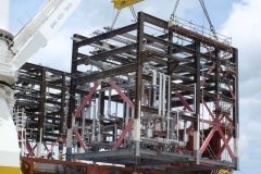 Crane lifting a large, rectangular steel structure with piping, likely an industrial module, outdoors.