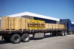 Flatbed truck loaded with wooden crates and yellow pallets, parked in front of a building under a clear blue sky.