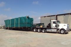 White semi-truck hauling a large, covered green object on a flatbed trailer; outdoors.