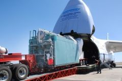 A large blue container being loaded onto a white aircraft from a red trailer at an airport on a clear day.