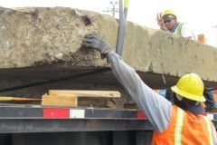 Workers loading a large, flat stone slab onto a flatbed truck. One worker reaches up, wearing a yellow helmet and orange vest.