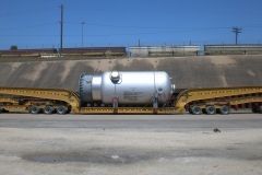 Large silver tank on a yellow, multi-wheeled transport vehicle, in an industrial setting.
