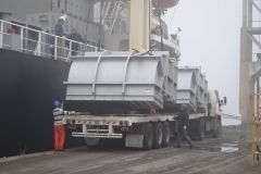 Truck with industrial equipment being loaded from a ship at a dock; workers in safety gear.