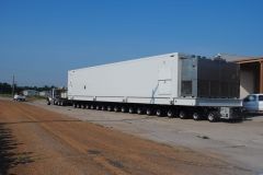 Large white container on a multi-wheeled trailer; truck on a dirt road; blue sky; sunny day.