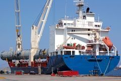 Blue and white cargo ship being loaded with a large cylindrical container by a crane at a dock.