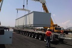 A crane lifts a large, gray modular structure over a multi-axle trailer on a paved area; workers in orange vests watch.