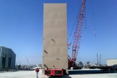 Tall beige concrete structure being lifted by a red crane outdoors, with a person walking in front.