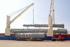 Cranes lifting and positioning large cylindrical industrial equipment onto flatbed trailers at a dock.