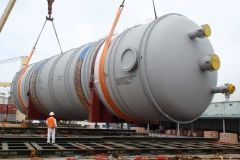 Large cylindrical industrial tank being lifted by crane, worker nearby. Gray, orange accents, outdoors.
