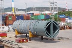 Large cylindrical industrial equipment in an outdoor setting, on yellow supports, with shipping containers in the background.