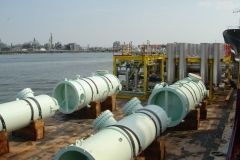 Several large, white cylindrical pipes on a barge, near a waterfront industrial area.