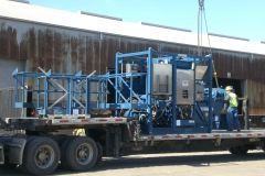 A blue industrial machine on a flatbed truck being unloaded, person in vest nearby.