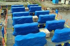 Blue-wrapped cargo on a metal deck. Boxes are secured and appear ready for transport, near water.