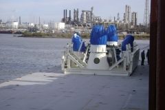 White machinery with blue-covered tops on a barge in front of an industrial area.