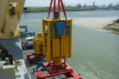 Yellow machinery being lifted from a ship, set against a water and industrial landscape.