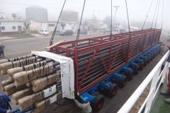 A large truck loaded with bundled pipes being lifted by cranes, outdoors on a foggy day.