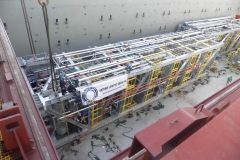 Large industrial machine being lifted by a crane on a ship; yellow and silver scaffolding, red and white ship.