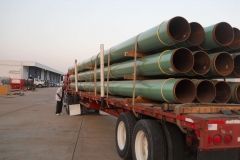 A truck bed loaded with green and brown pipes; a person stands nearby at a loading dock.