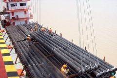 Workers loading long black pipes onto a barge at a dock.