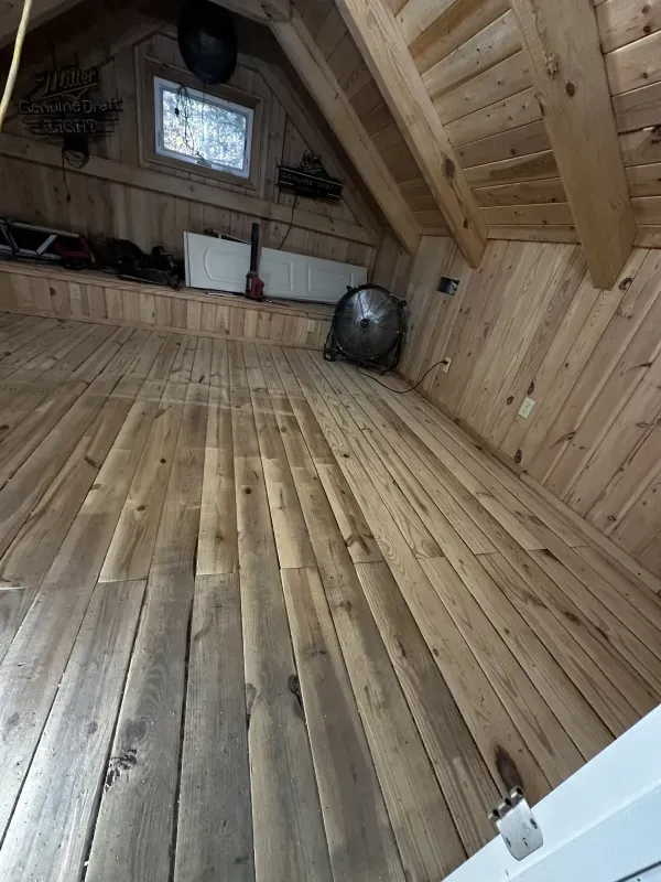 A wooden floor in an attic with a fan and a window.