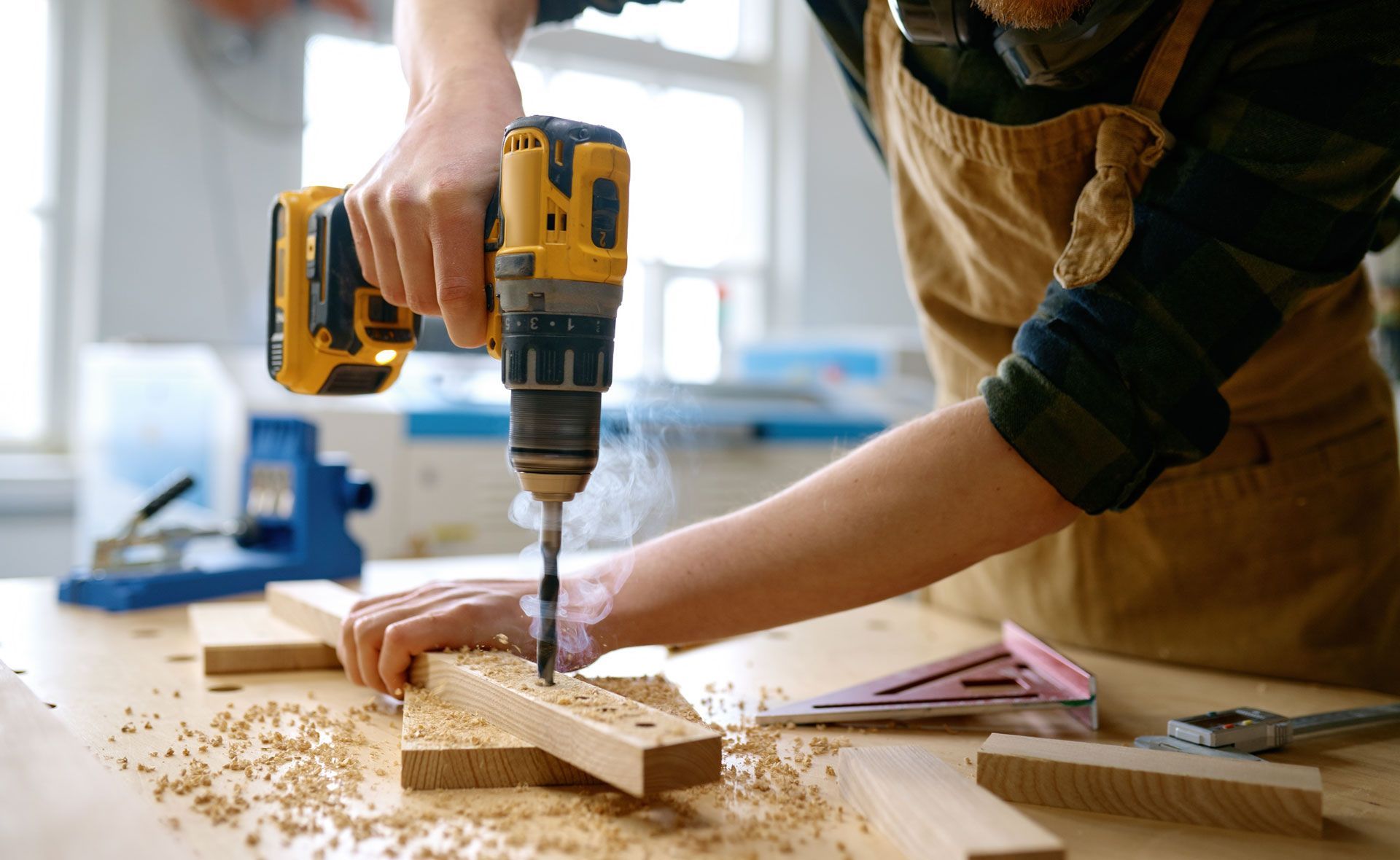 A person wearing an apron uses a yellow power drill to bore a hole into a wooden board, with smoke rising from the drill.