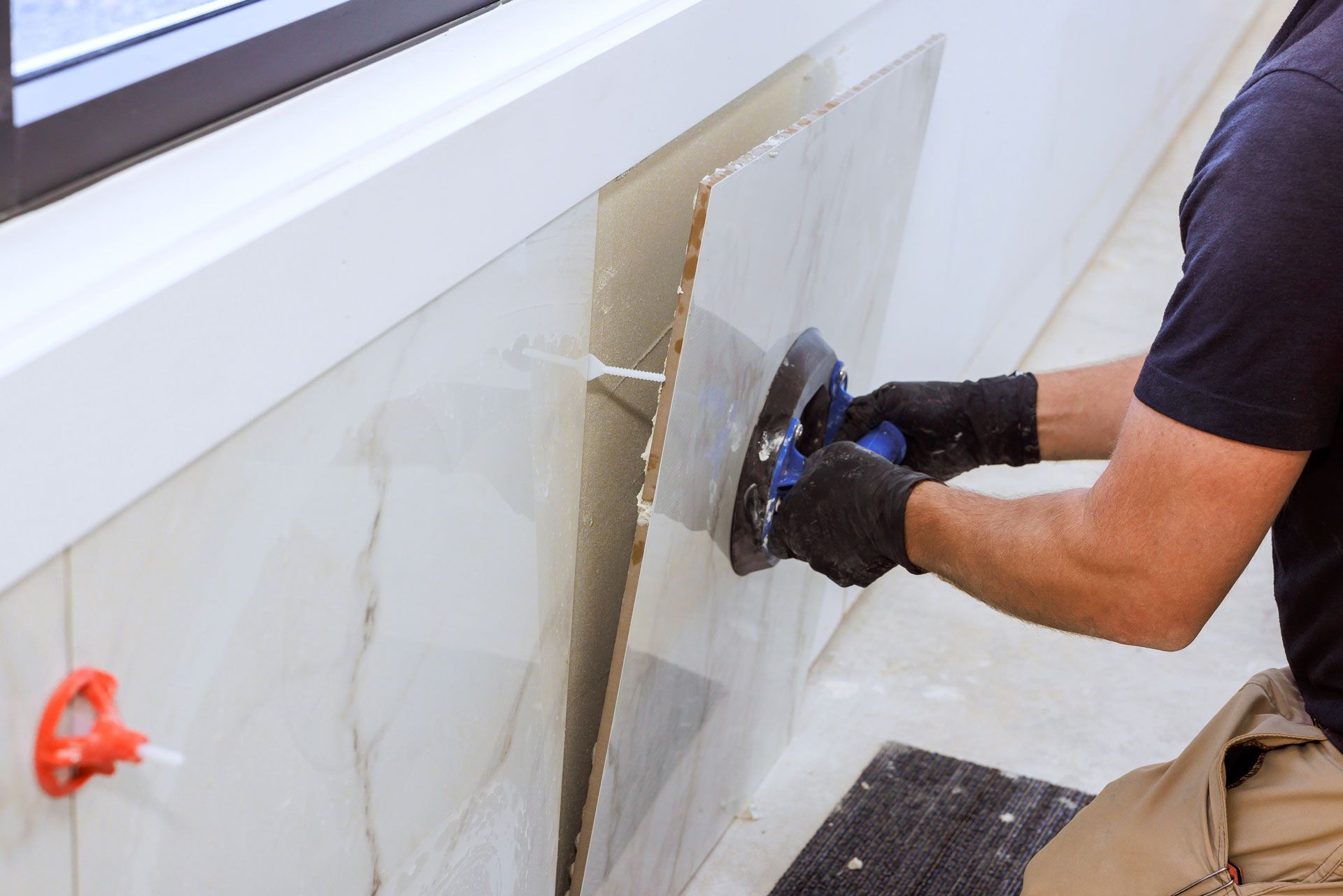 A worker in black gloves uses a suction handle to install a large, light-colored marble-patterned tile on a wall.